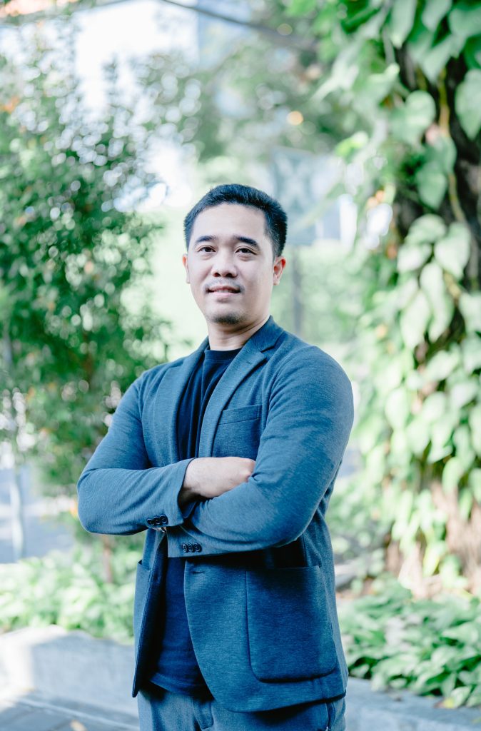 Arekha stands outside with foliage behind him smiling and wearing a grey suit jacket.